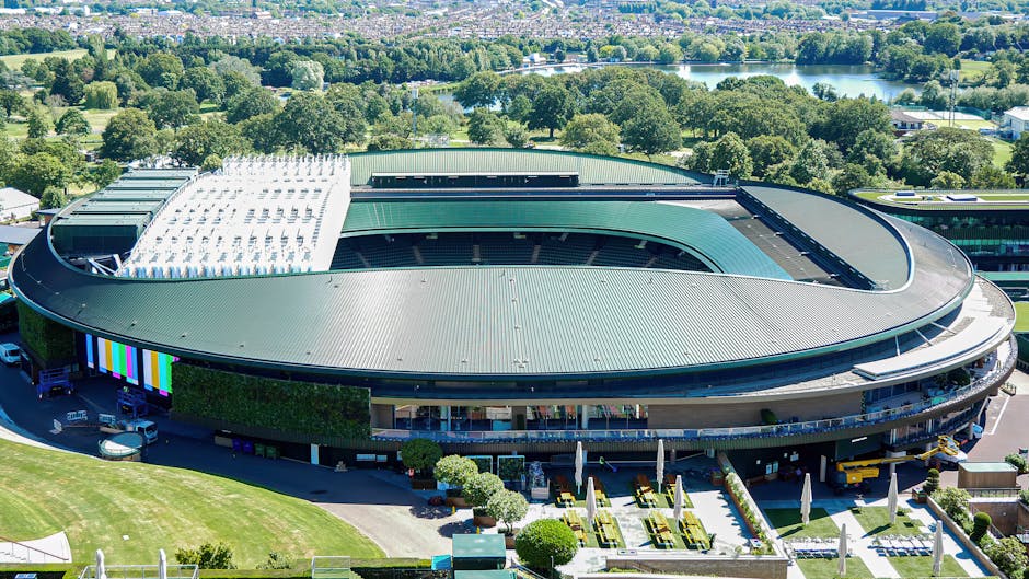 An aerial view of Twickenham Stadium showcasing its large, oval-shaped roof with a section featuring white solar panels. The stadium is surrounded by green trees and open fields, with a parking area a