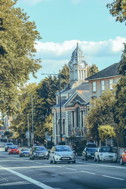 A street scene in a residential area with a historic clock tower featuring a pointed spire and a large clock face, situated behind a row of mature trees with green foliage. The street is populated wit